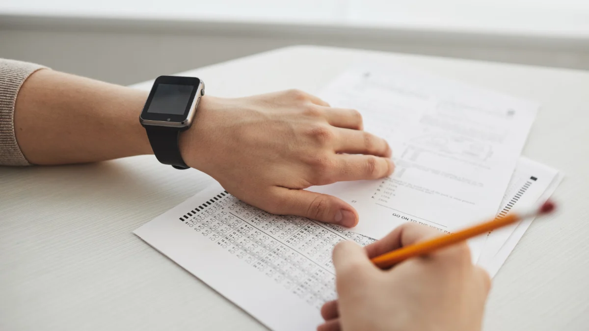 Edukator starannie sprawdza arkusze egzaminacyjne przy użyciu czerwonego markera przy biurku, ilustrując skoncentrowane ocenianie egzaminu