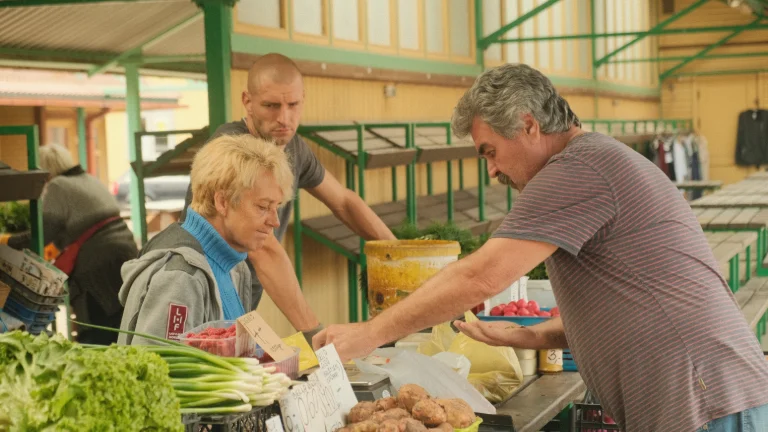 Scena targu rolniczego z wielu sprzedawcami oferującymi świeże warzywa i ziemniaki, w tle stojące stände i ludzie kupujący